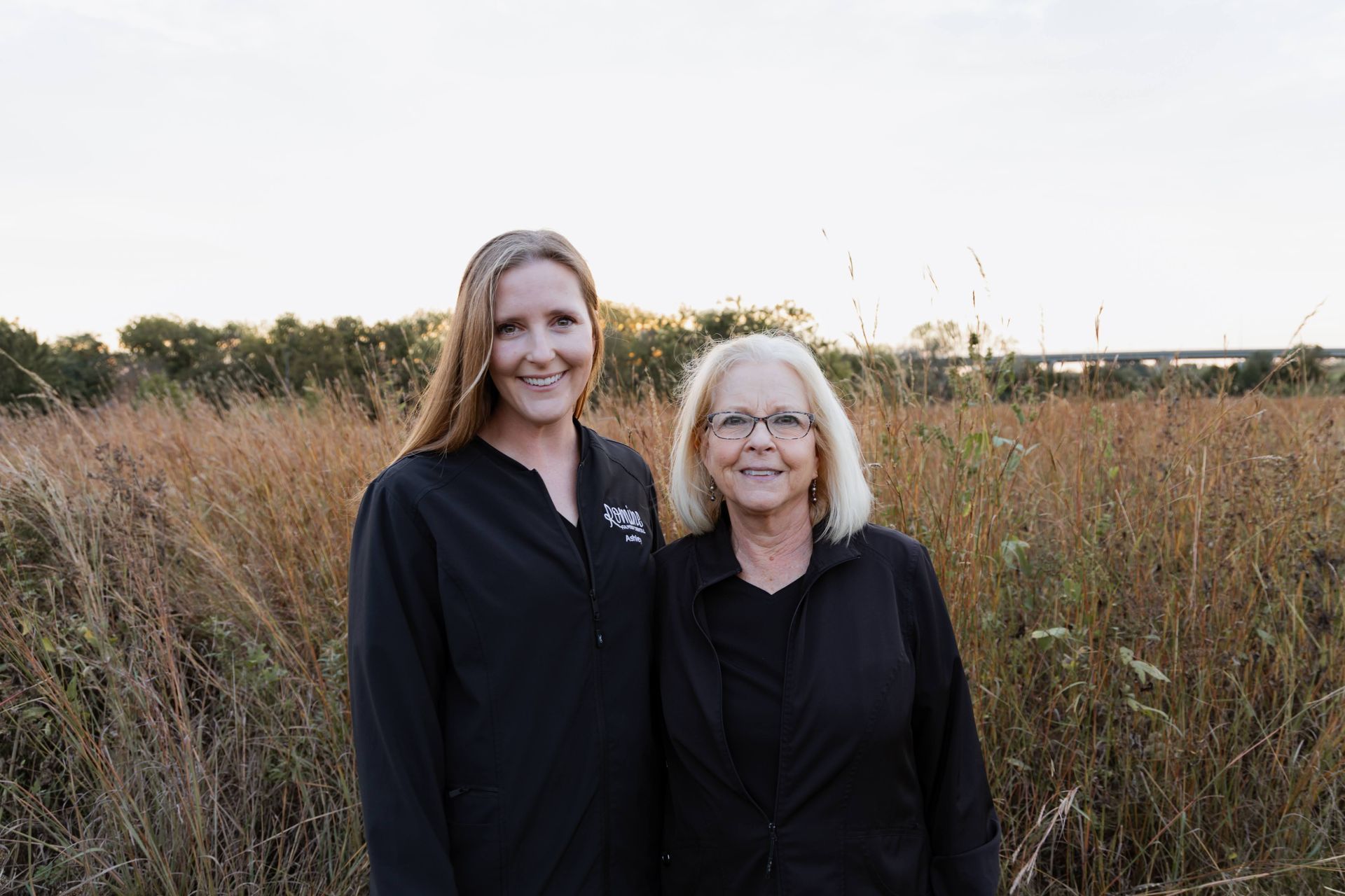 Two women stand in tall grass field, both wearing black. One has long hair, and the other has glasses.