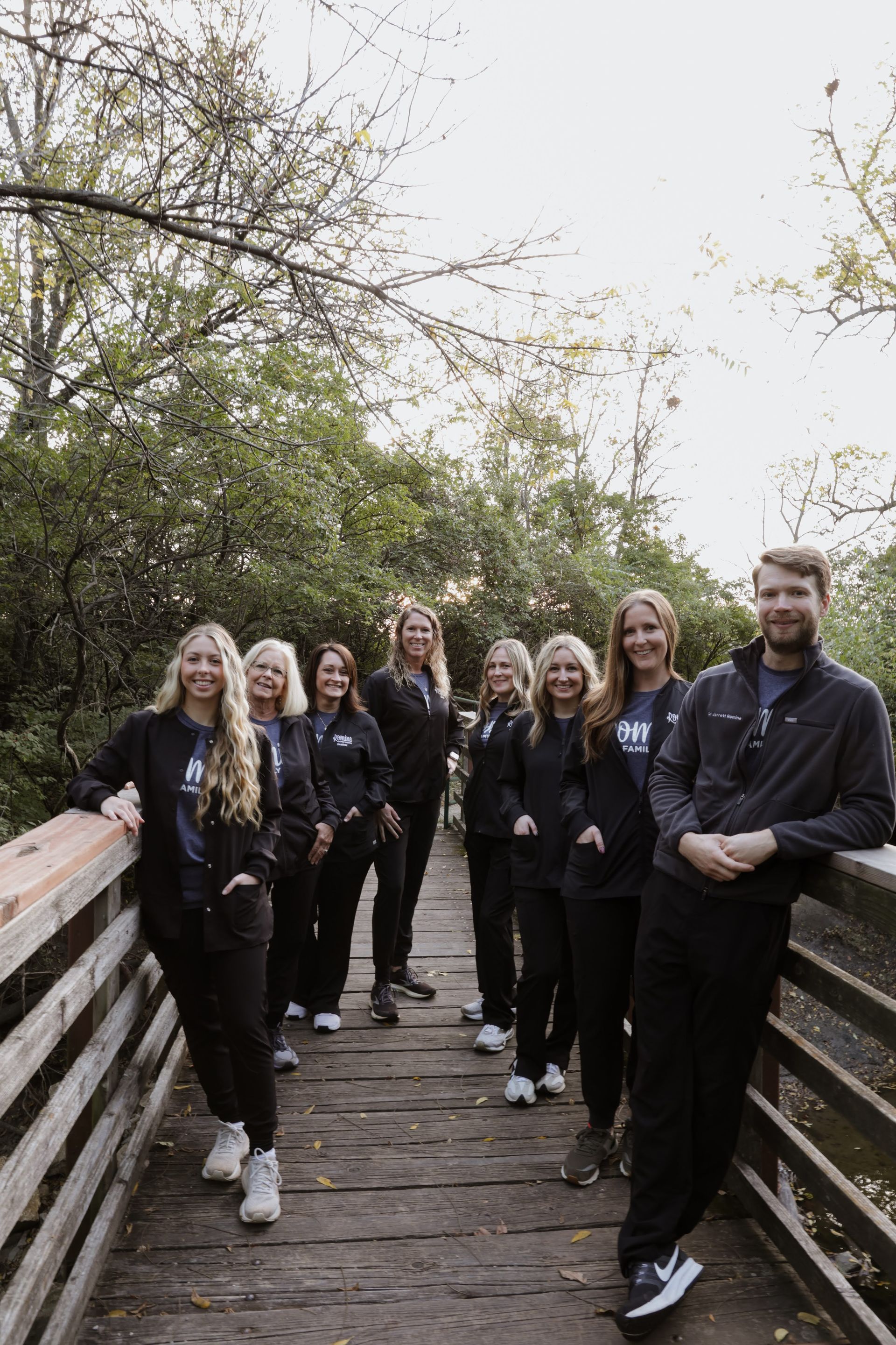 Group of people in black outfits on a wooden bridge in a natural setting, smiling.