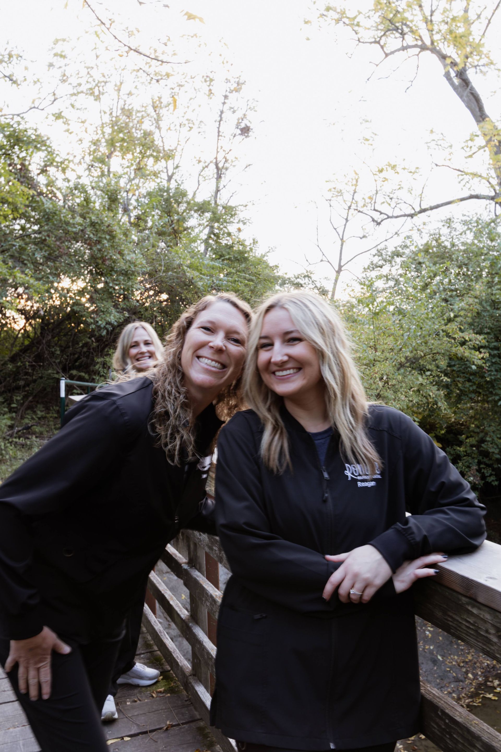 Three women smiling on a wooden bridge in a wooded area. The two in front are wearing black jackets.