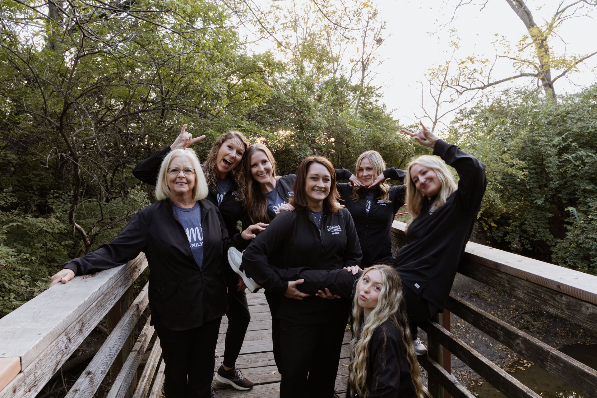 Group of eight people on a wooden bridge, posing with various hand gestures and smiles in a wooded area.
