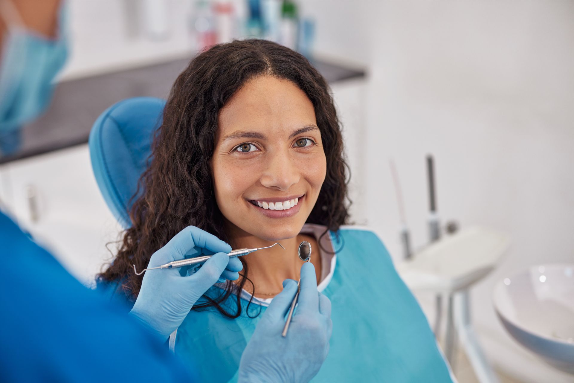 A dentist wearing blue gloves performs an exam on a smiling patient in a bright, modern dental office.