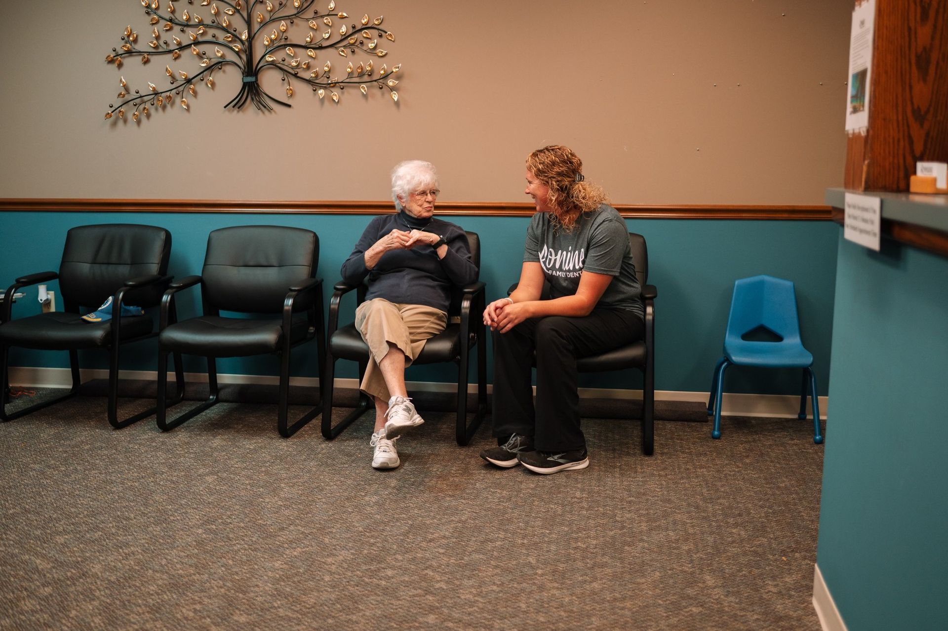 Two people in a waiting room, one gesturing. Dark chairs line the wall, a tree art piece hangs above.