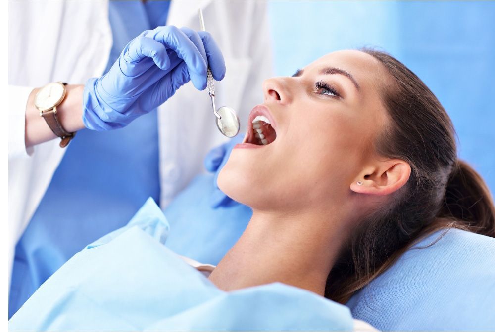 Woman at the dentist with mouth open; doctor in blue gloves examines teeth with a mirror.