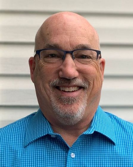 A bald man with glasses and a beard is wearing a blue shirt and smiling.