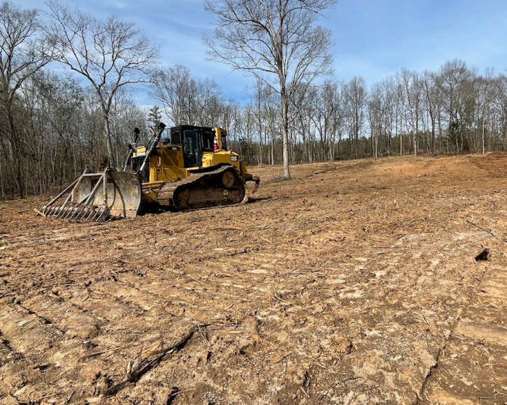 A bulldozer is driving through a dirt field with trees in the background.