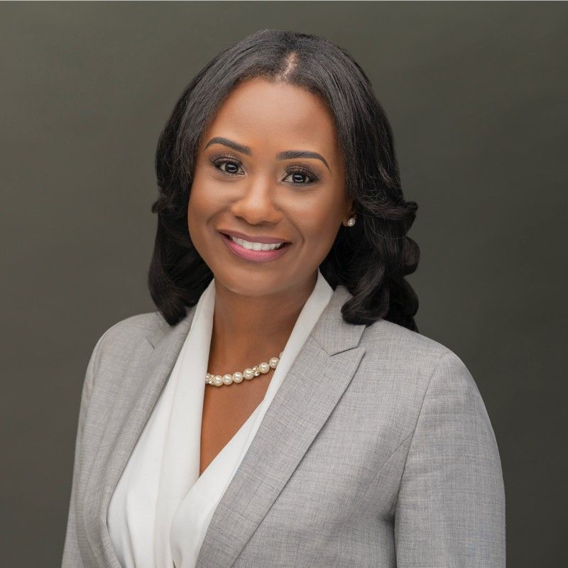 Woman in a gray suit and pearl necklace smiles at the camera against a gray background.