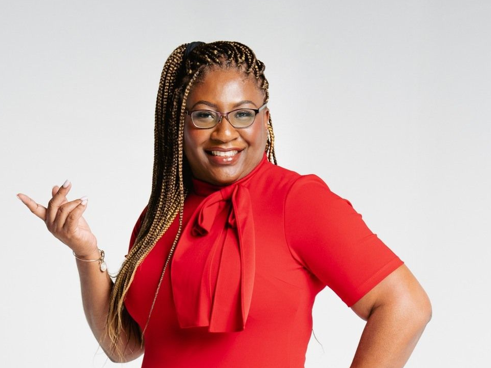 Woman with long, braided hair, wearing glasses and a red top, smiling and gesturing.