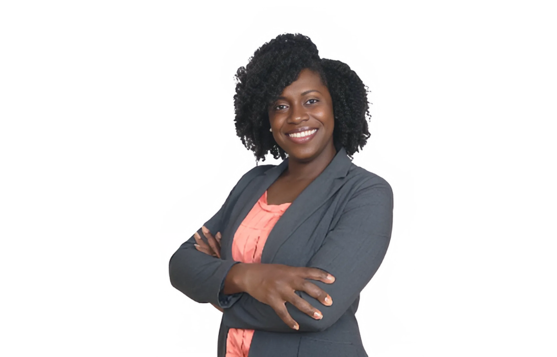 Black woman with arms crossed, smiling, wearing a grey blazer and peach top.
