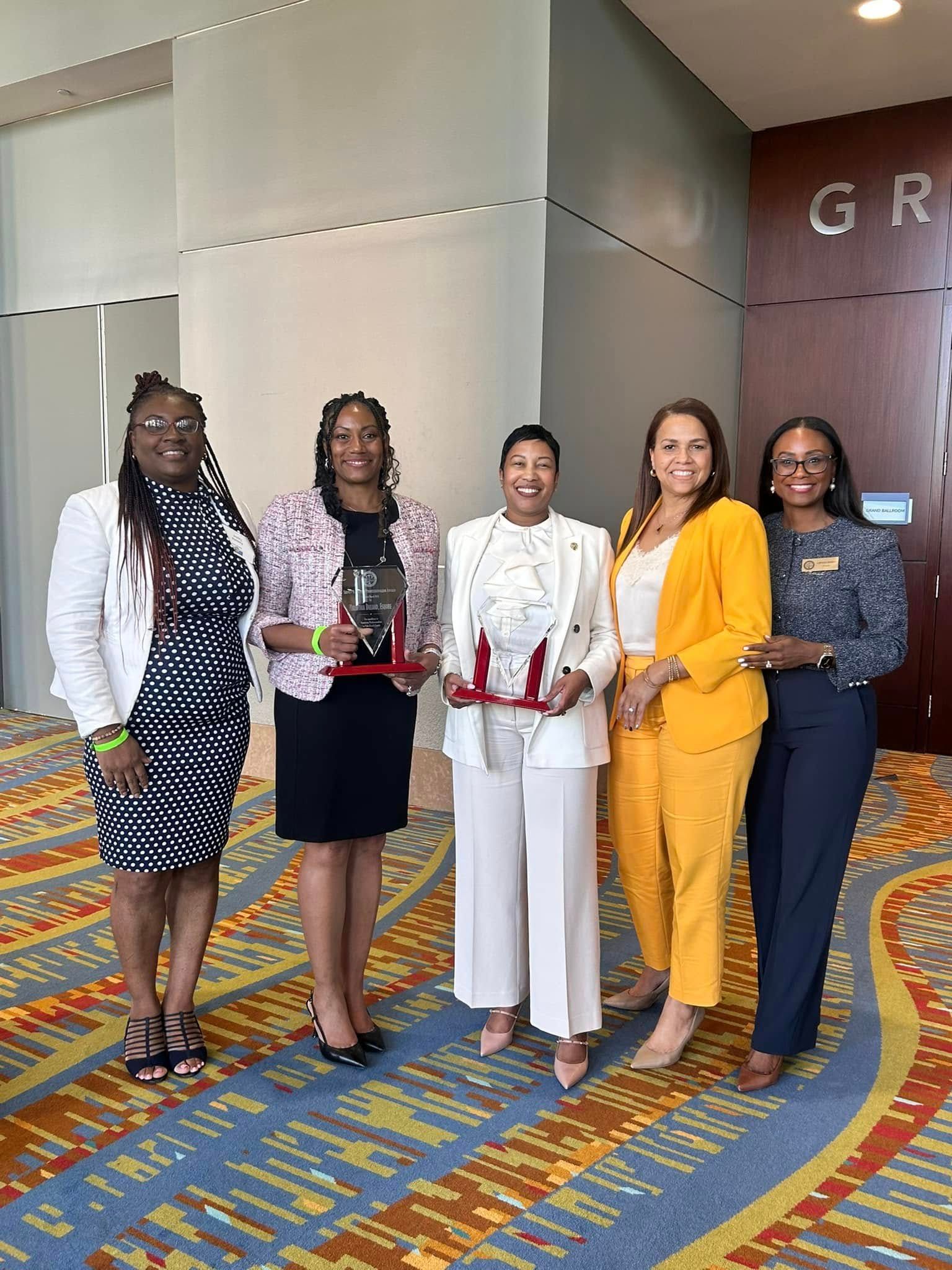 Five women in business attire, holding awards, smiling, in a building with