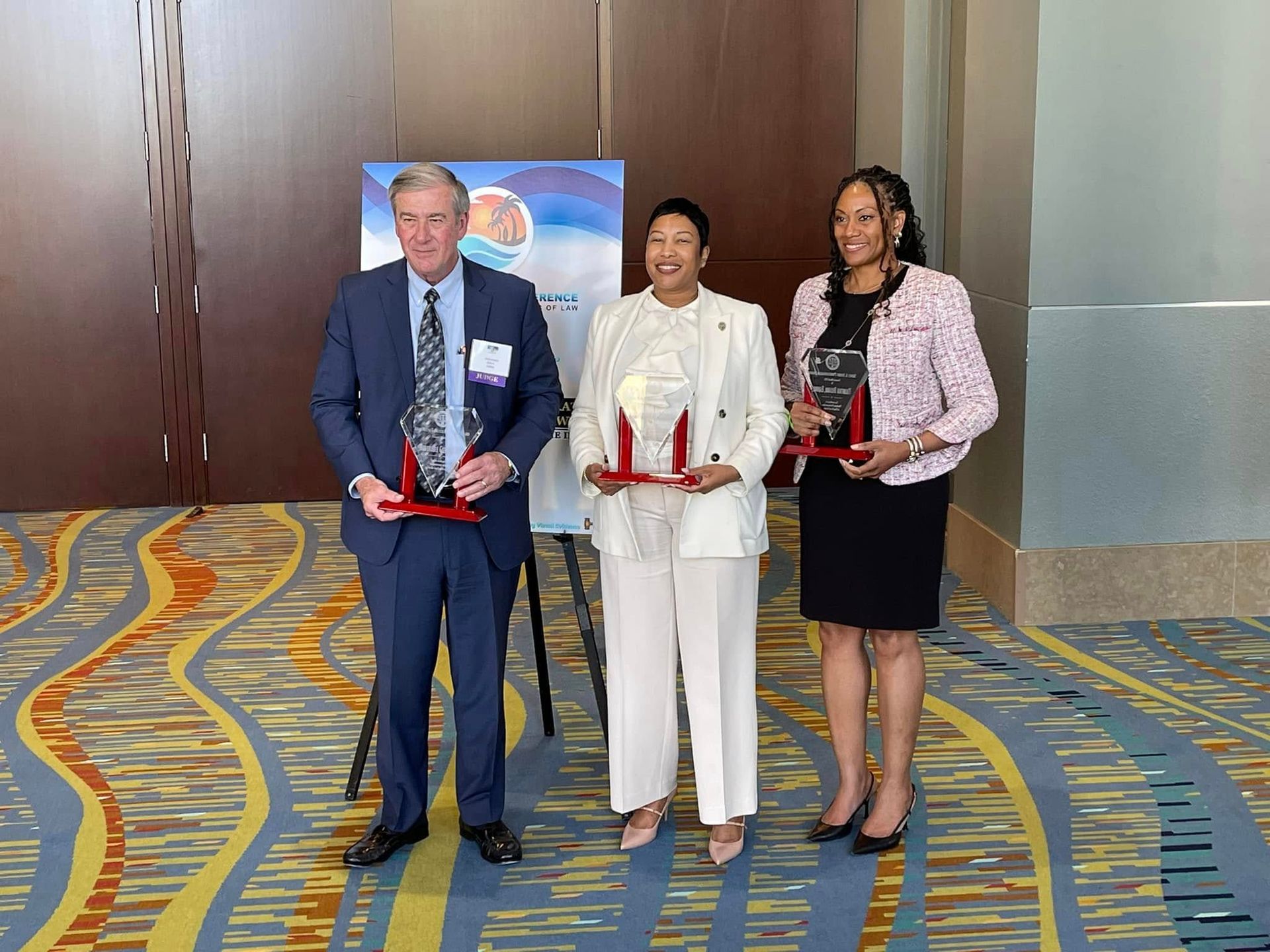 Three people holding awards, posing for a photo. Man in suit, woman in white, and woman in pink jacket.