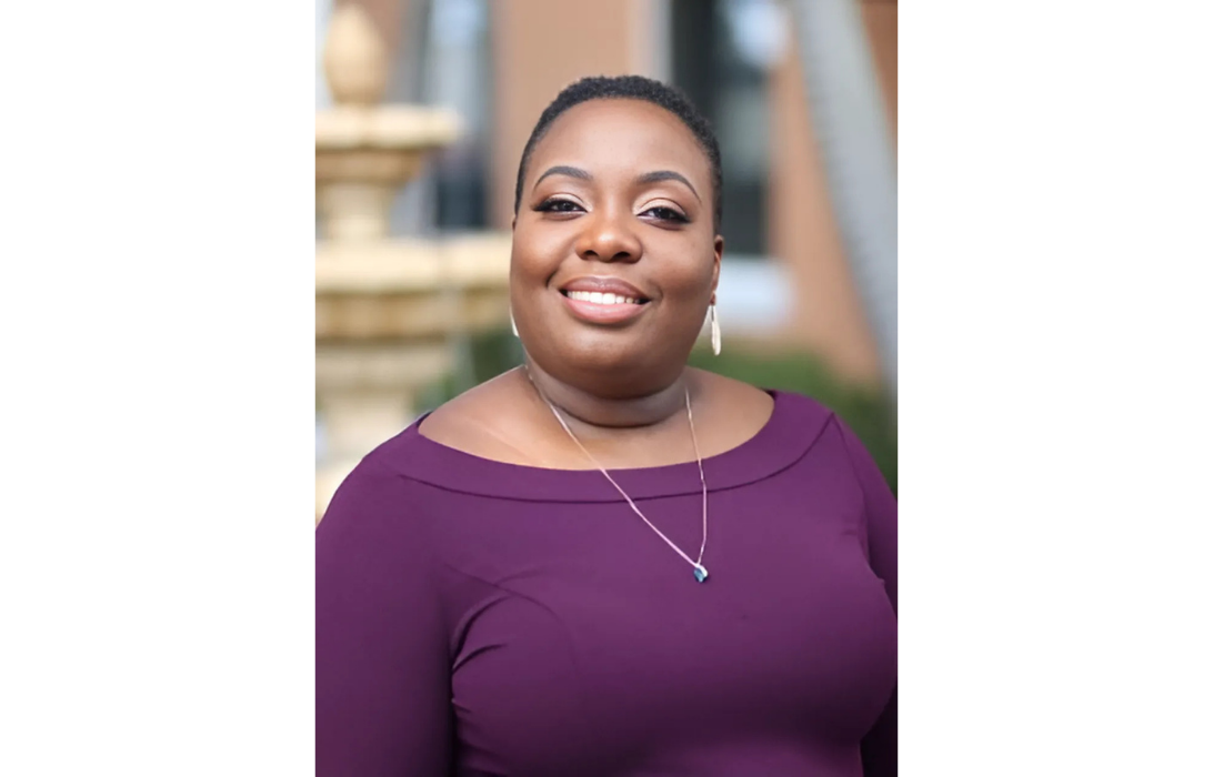 Woman with dark skin smiles, wearing a purple off-the-shoulder top, standing outside near a fountain.