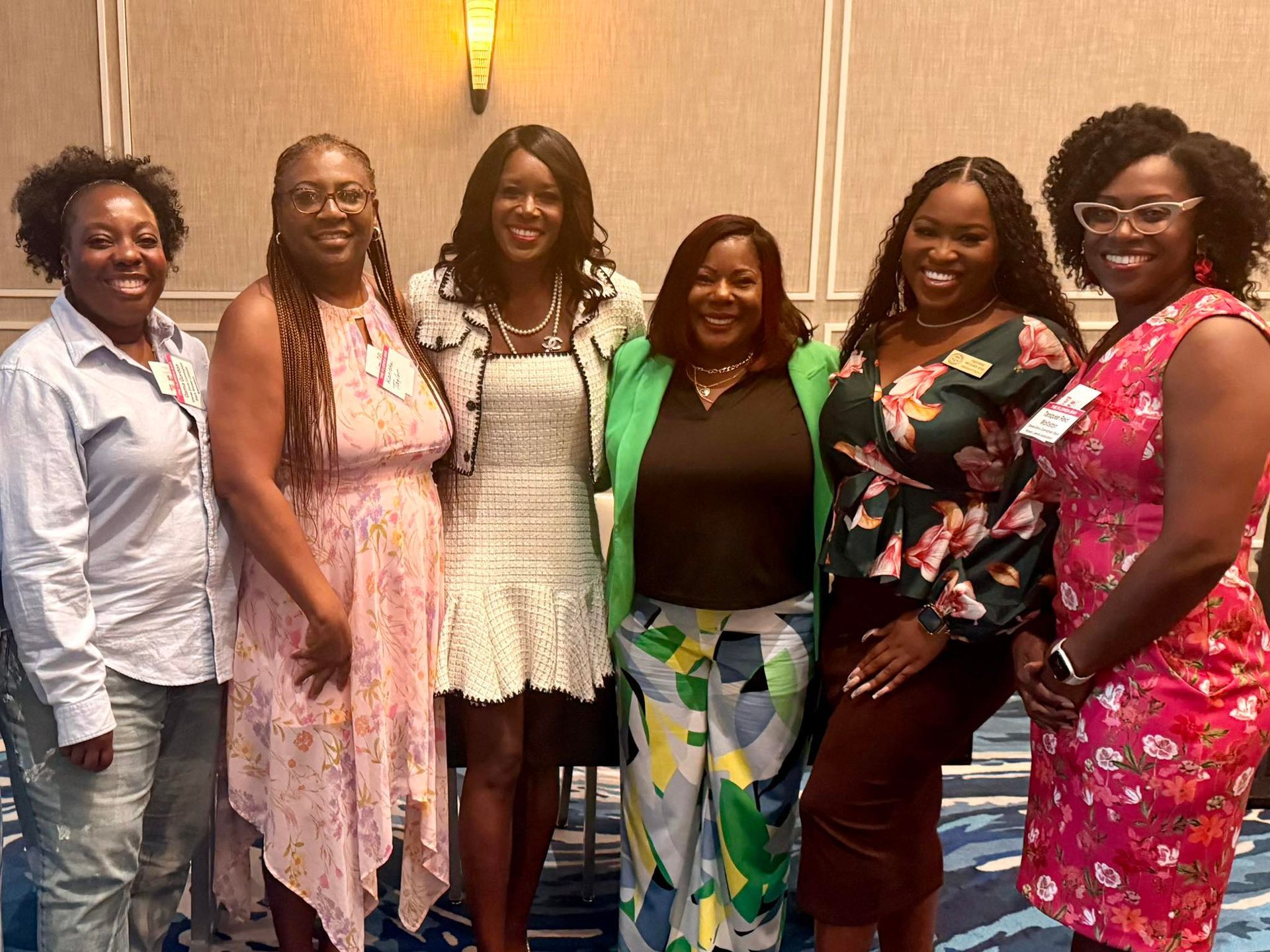 Six smiling women pose together indoors. They're dressed in various colorful outfits, including dresses, pantsuits, and floral tops.