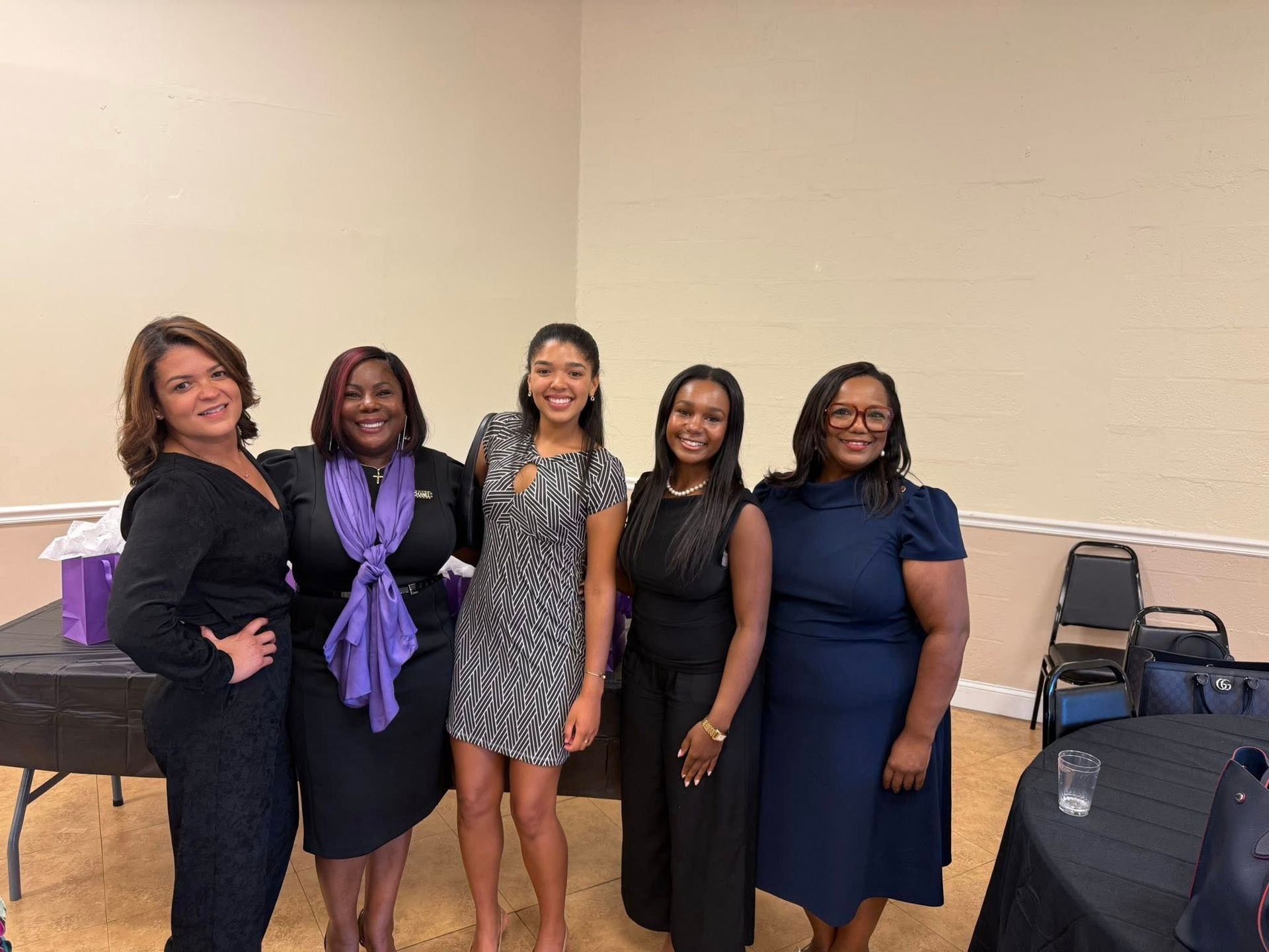 Five women posing together at an event, indoors. They are dressed in business attire, smiling.