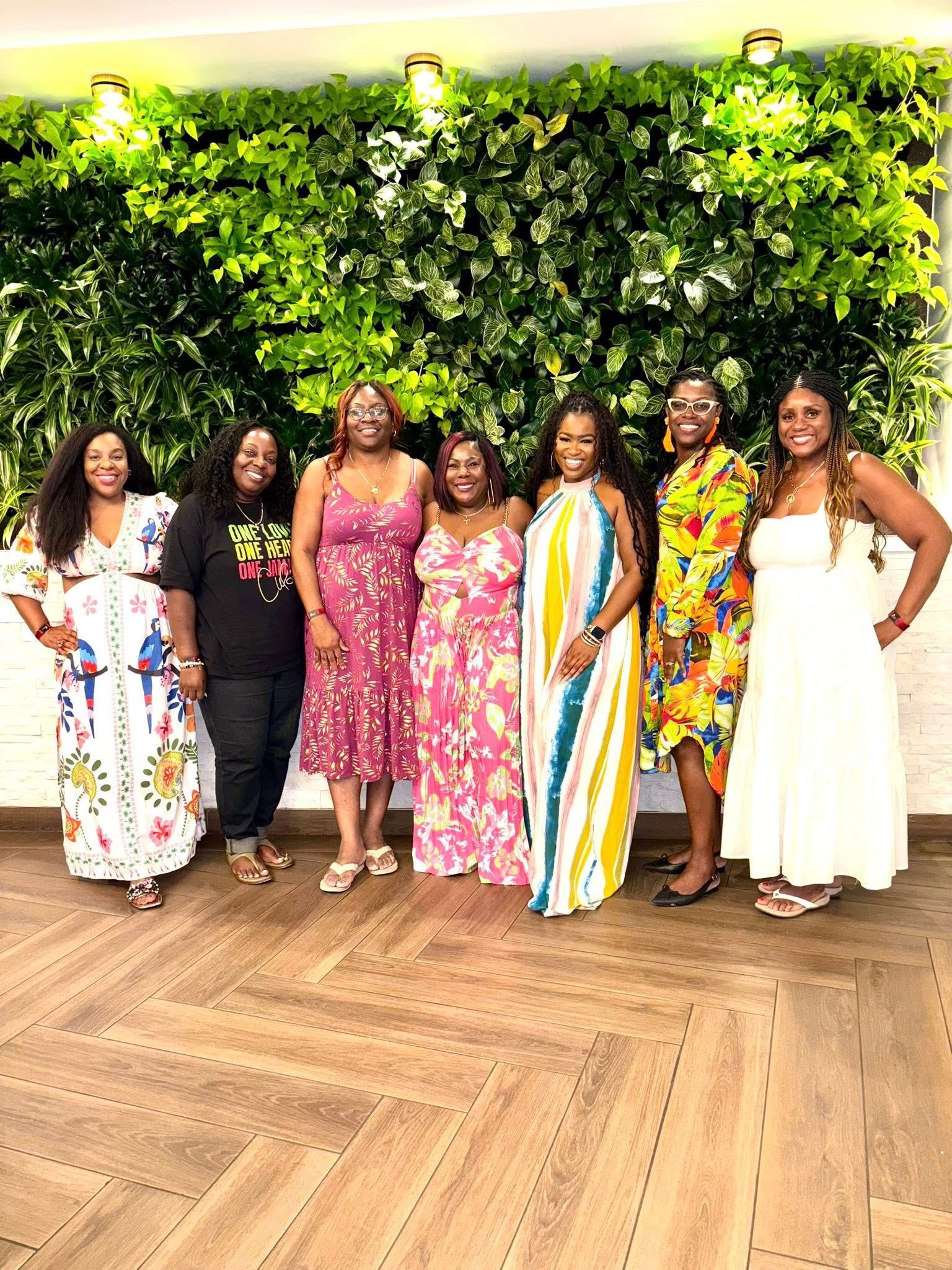 Seven smiling women in colorful dresses pose in front of a green wall.