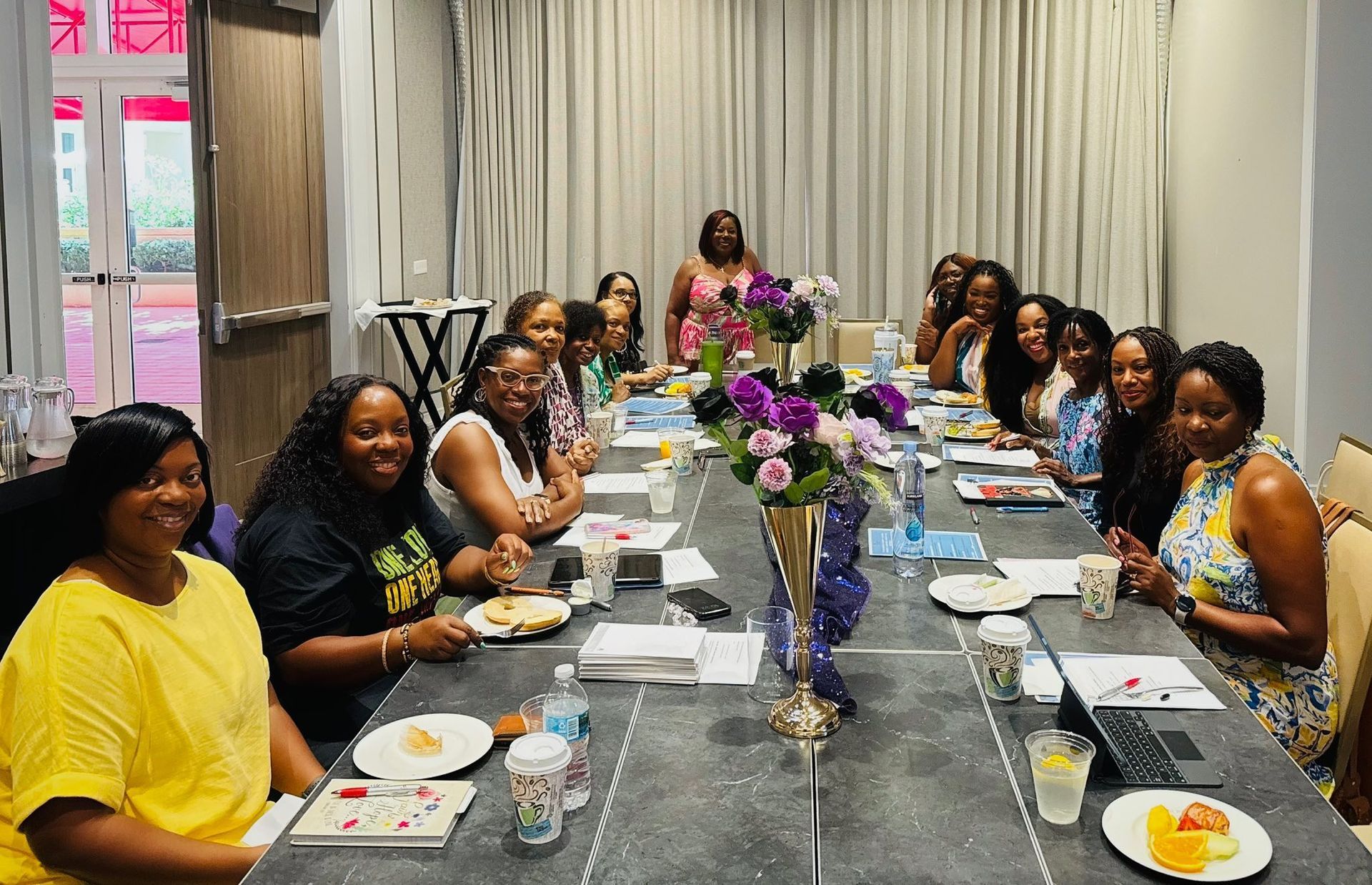 Group of women at a table, smiling at the camera. Flowers and food are on the table.