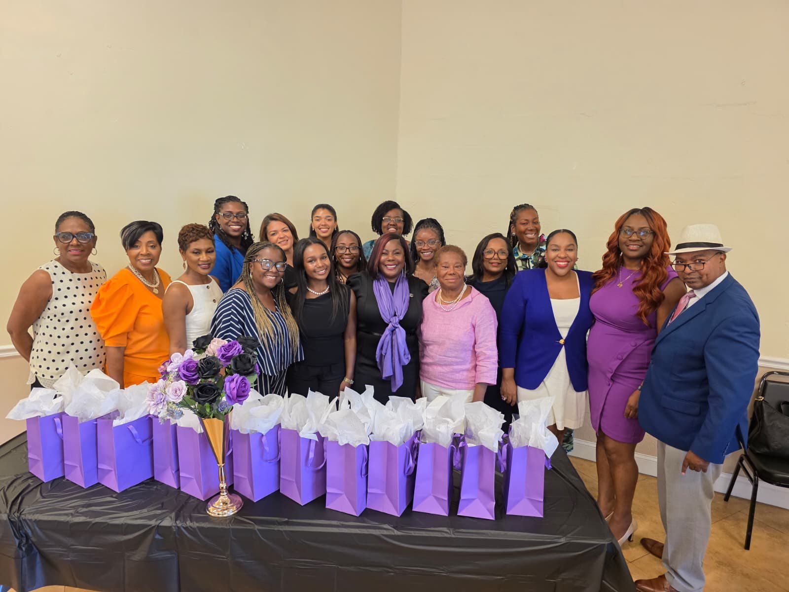 Group of people pose with gifts at a celebration; women in various colored outfits and a man in a blue blazer and fedora.