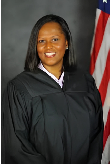 A Black woman judge in a black robe smiles, with the American flag behind her.
