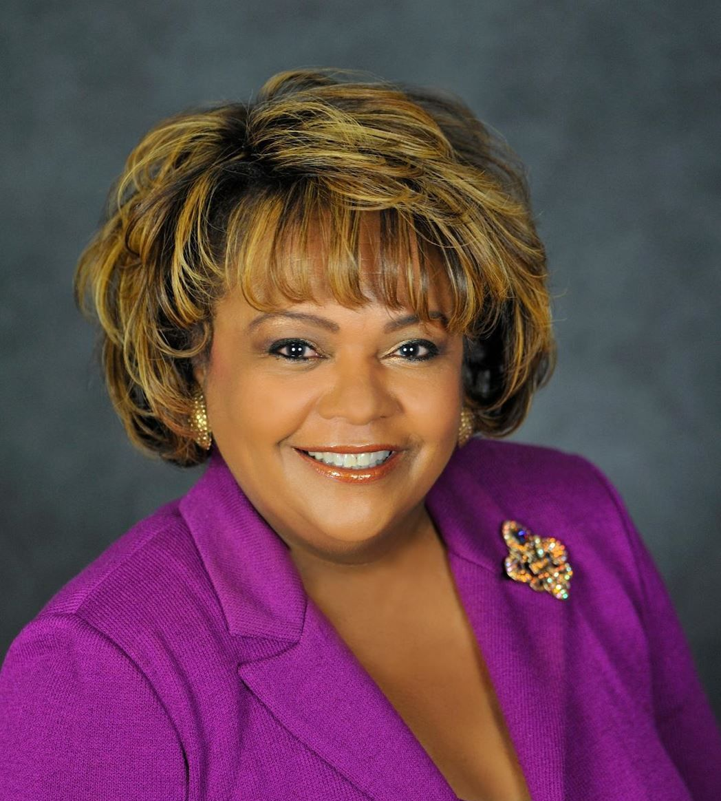 Woman with brown hair smiling, wearing a purple blazer, and a brooch.