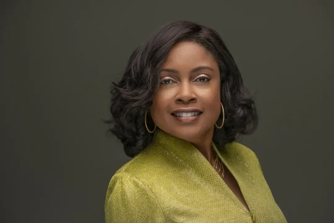 Woman with dark hair smiles, wearing a yellow jacket and gold hoop earrings against a gray background.