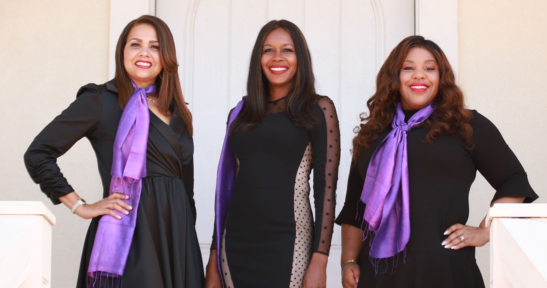 Three women in black dresses with purple scarves, posing outside.