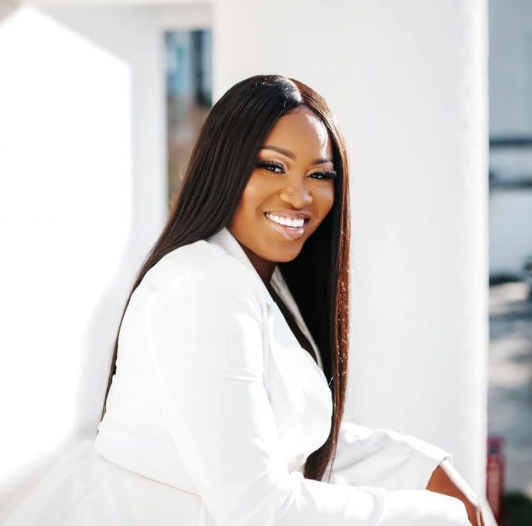 Woman in white blazer smiles leaning against a white pillar.
