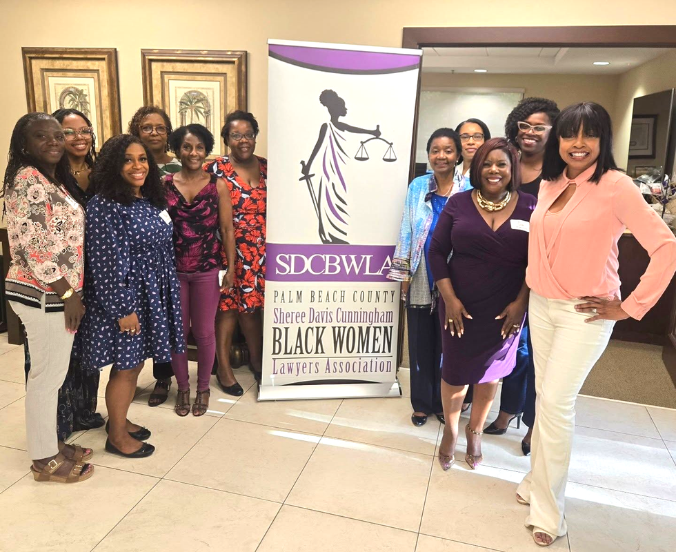 Group of Black women pose in front of a banner with a logo. Hallway setting.