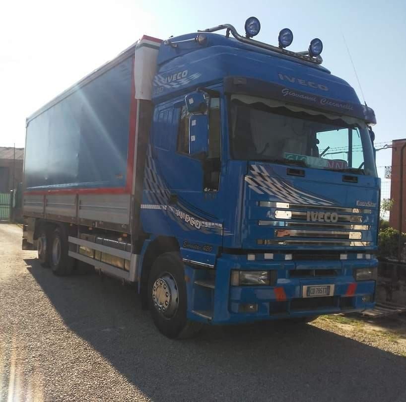 Camion Iveco blu parcheggiato fuori. Rimorchio furgonato e cabina con luci sul tetto. Giornata di sole.