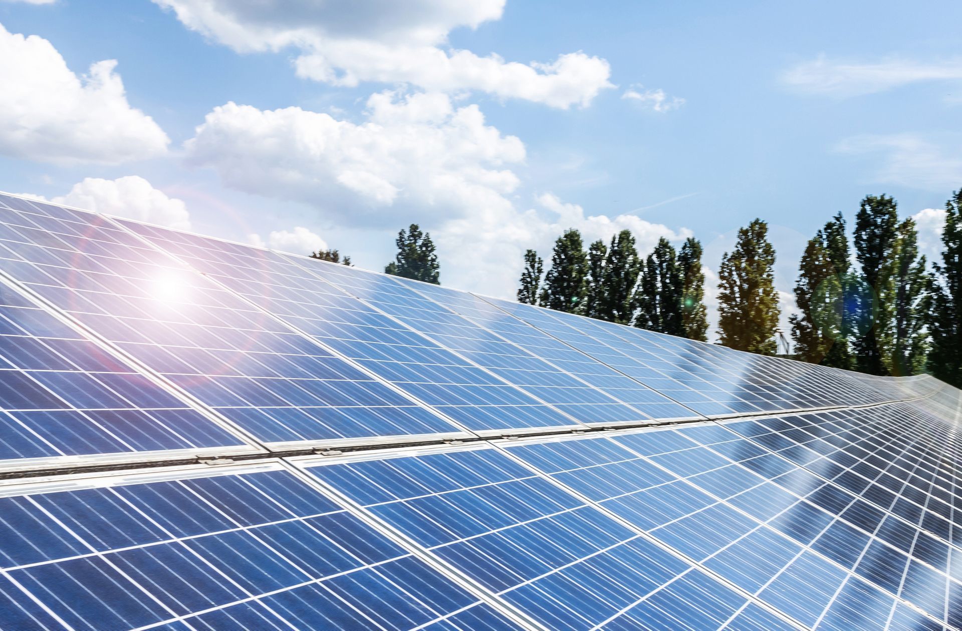 Pannelli solari, blu, con la luce del sole che si riflette su di essi, contro un cielo azzurro brillante con qualche nuvola e qualche albero.