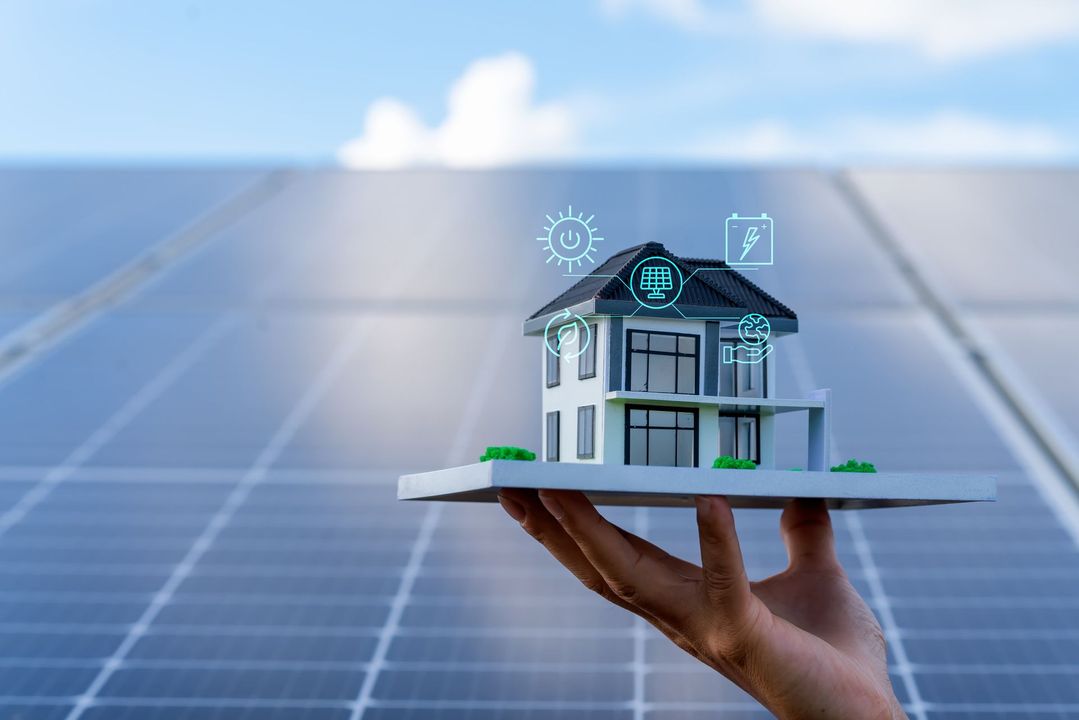 Mano che regge un modellino di casa con pannelli solari sullo sfondo; icone della tecnologia per la casa intelligente.