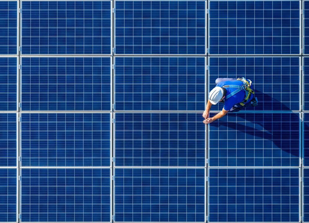 Operaio con dispositivi di sicurezza sui pannelli solari, ispeziona l'installazione. Vista aerea, blu e bianca.