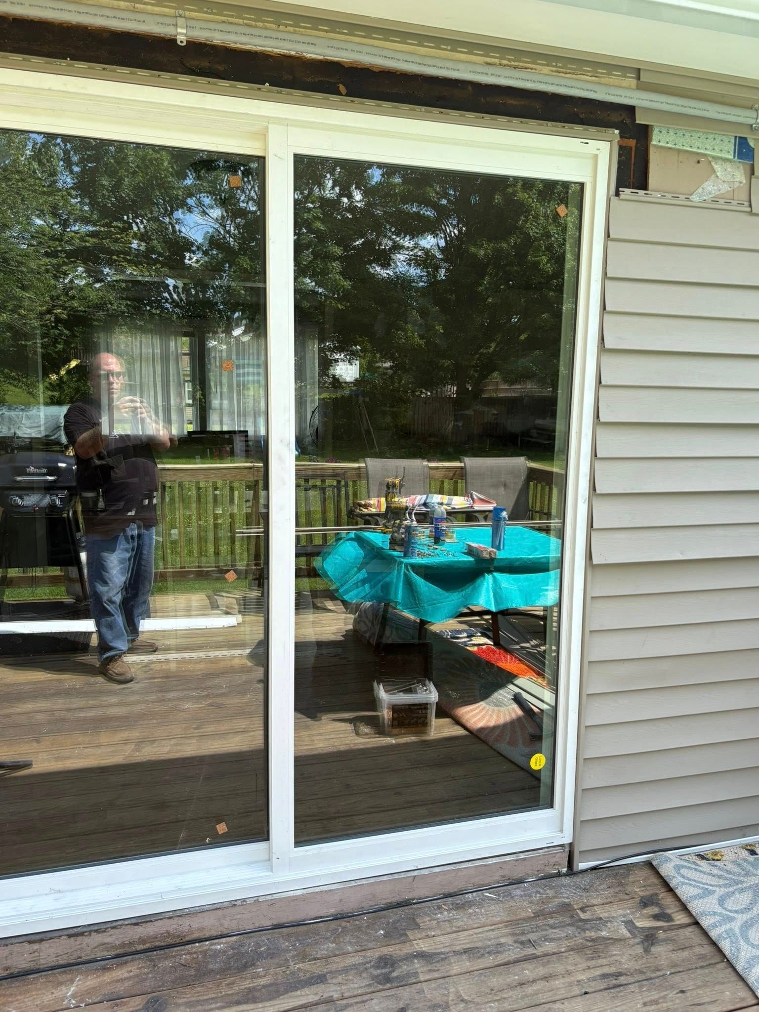 Sliding glass door reflects a person, deck, and outdoor table with blue tablecloth. Beige siding.