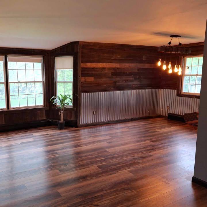 Empty living room with wood floors, walls, and tin wainscoting. Windows and a light fixture are visible.