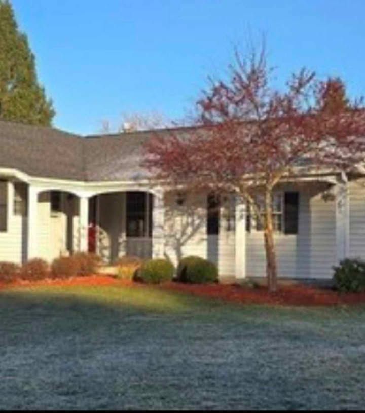 White house with porch, red-leafed tree, green lawn, clear blue sky.