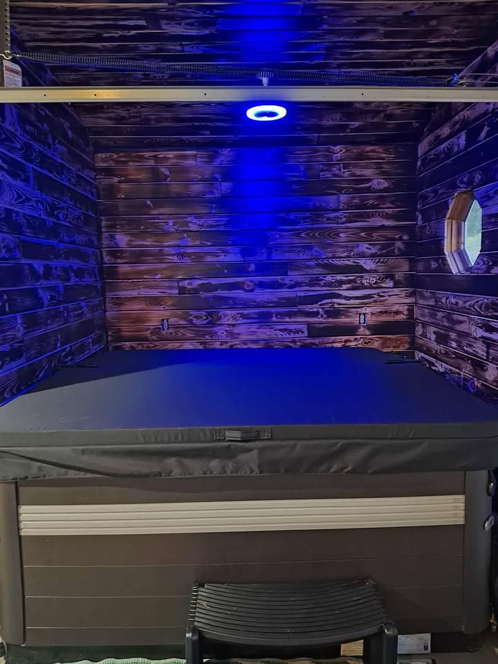 A hot tub under blue light within a rustic wood-paneled enclosure; a small, round window is visible.