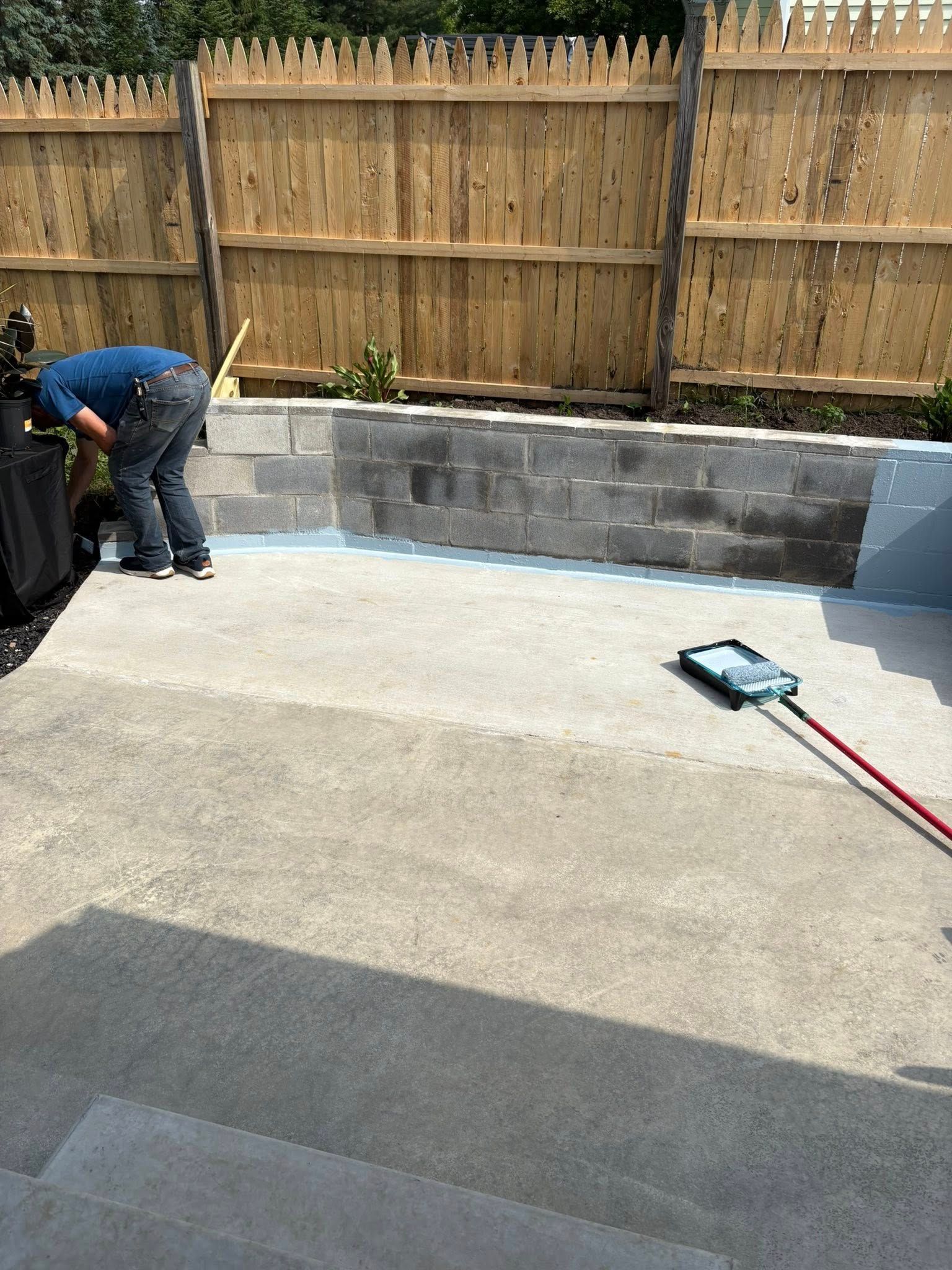Person painting a concrete patio, with a cinder block wall and wooden fence in the background.