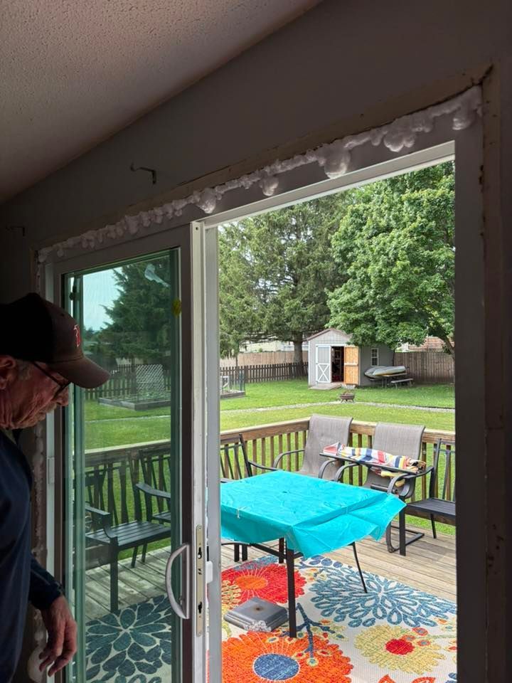 Person looking at a sliding glass door open to a backyard deck with a table, chairs, and shed.