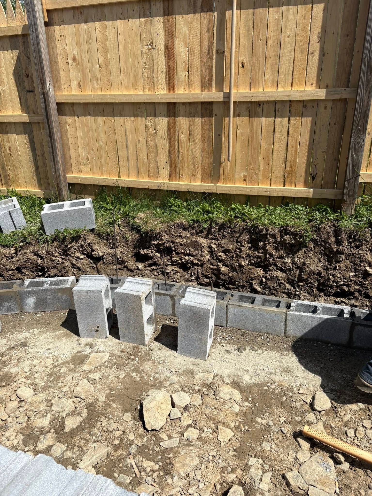 Concrete blocks are arranged along a dirt strip in front of a wooden fence, likely for a garden bed.
