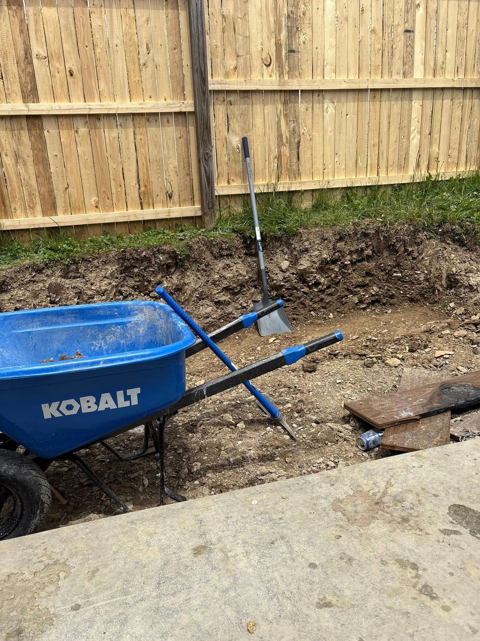 Blue wheelbarrow next to a dirt trench with a shovel, near a wooden fence.