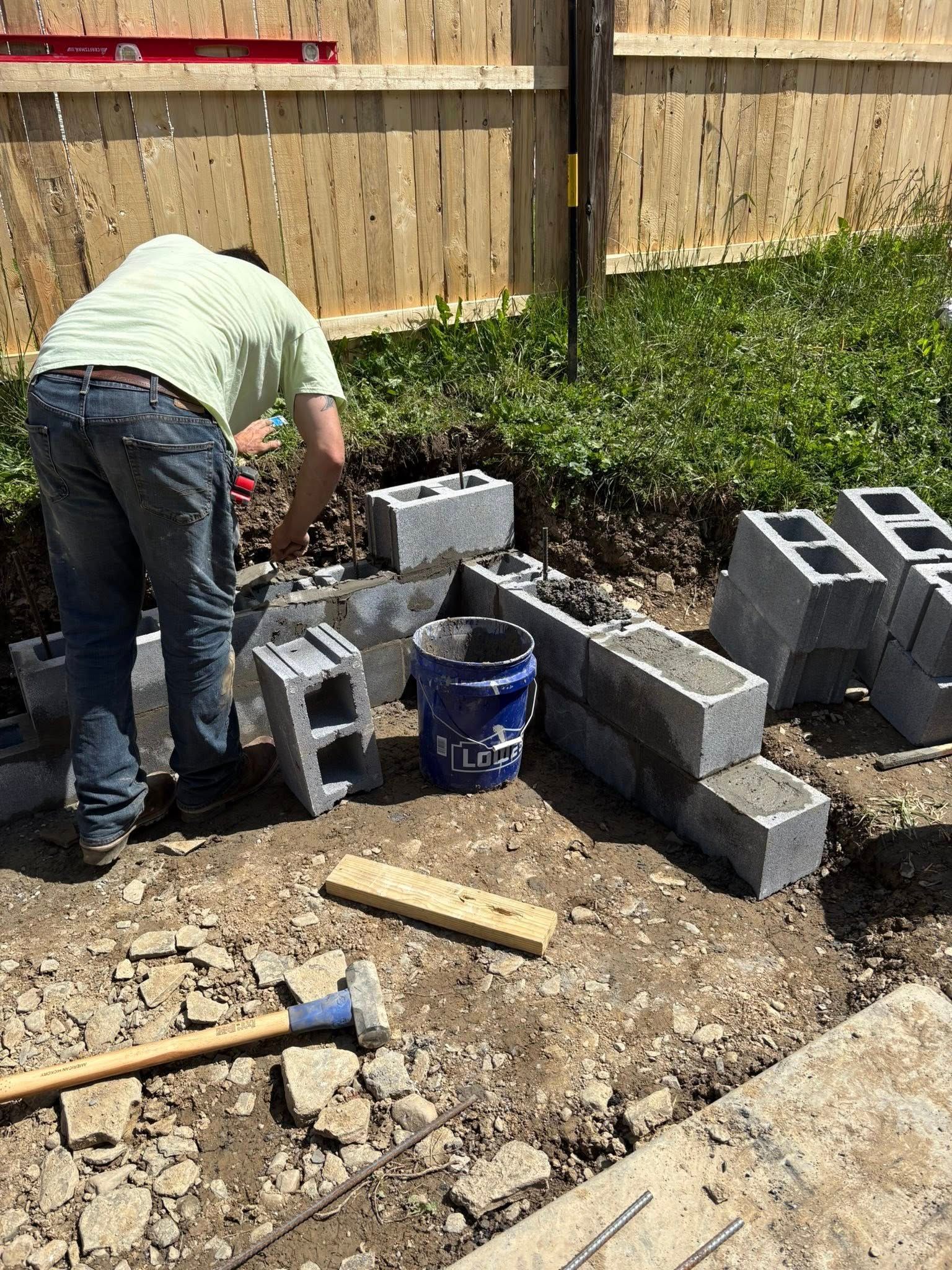 Person laying cinder blocks next to a fence. Blocks, bucket, hammer, and level are visible.