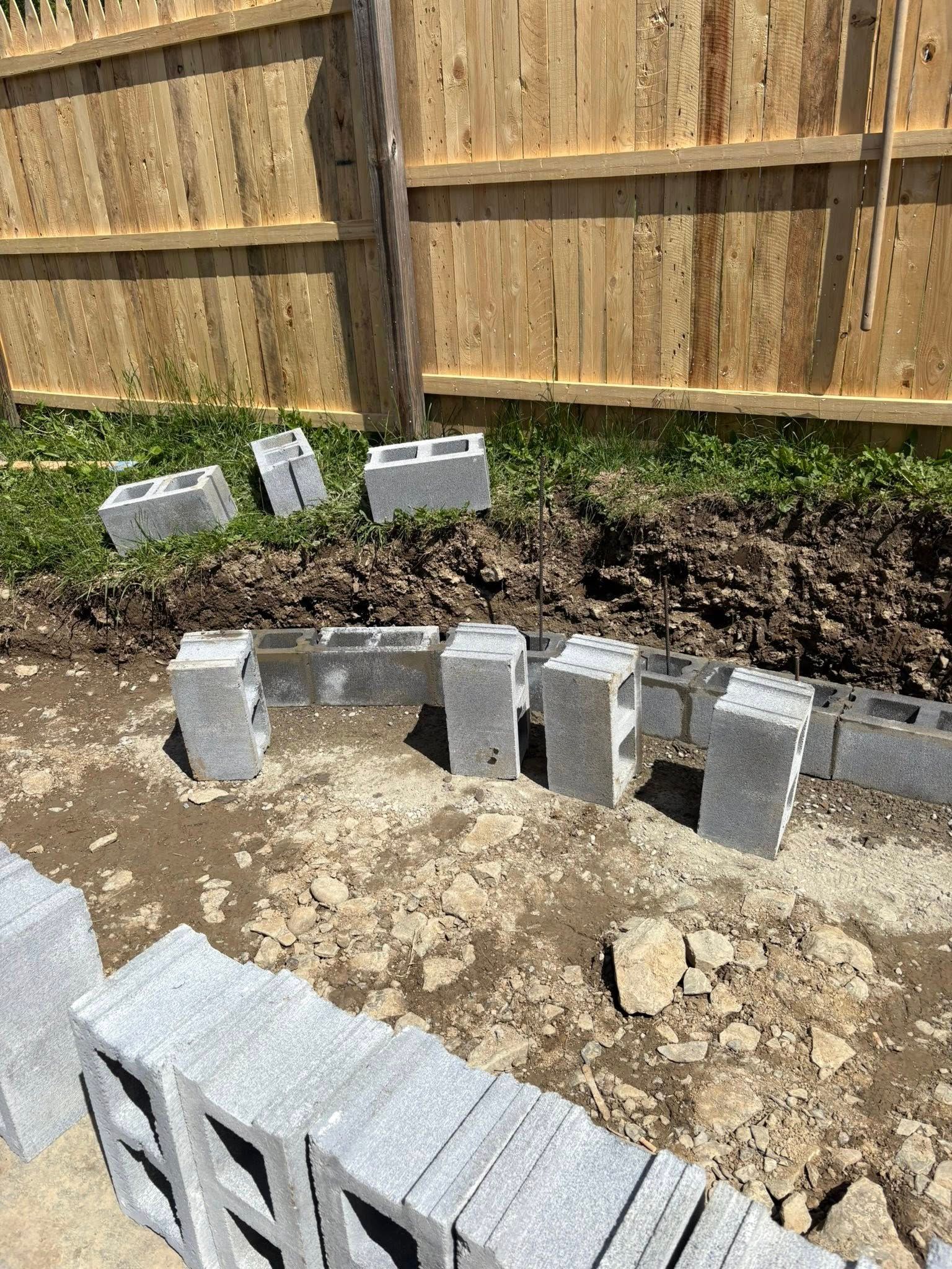 Concrete blocks arranged along a muddy area near a wooden fence, possibly for construction of a raised garden bed.