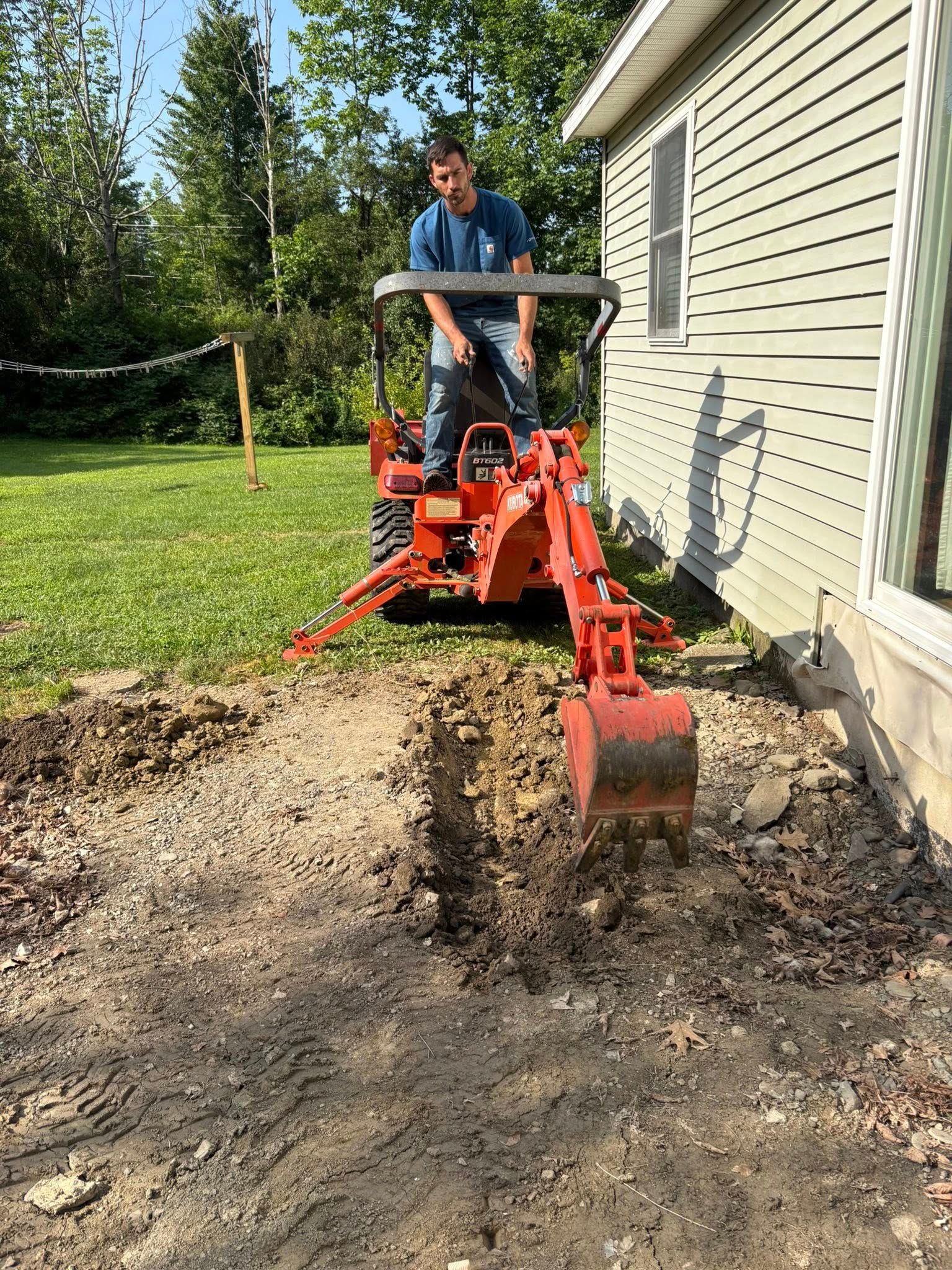 Man operating an orange backhoe digging near a house. Sunny day, grassy yard.