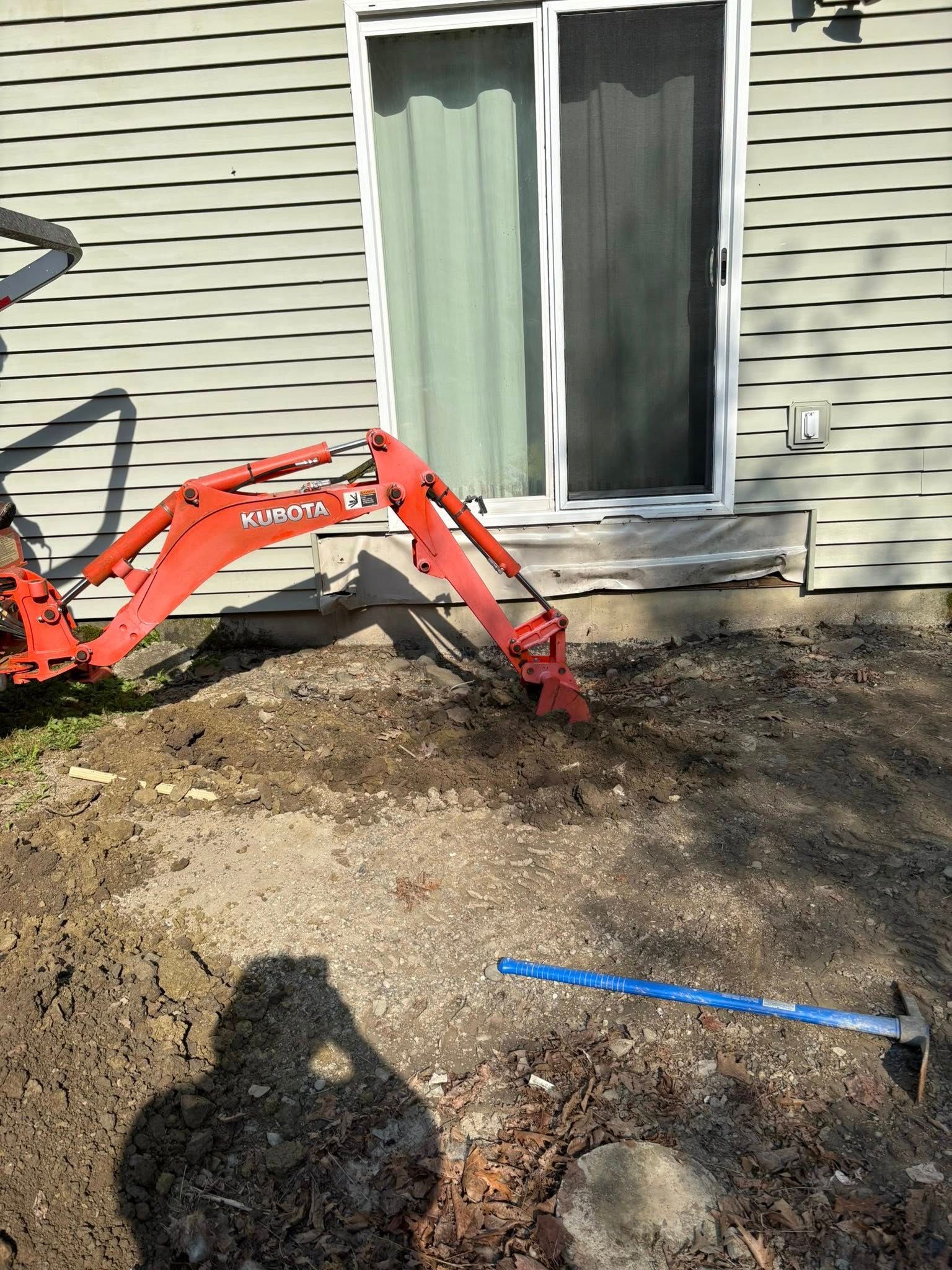 Red mini excavator digging dirt near a sliding glass door and a blue hoe.