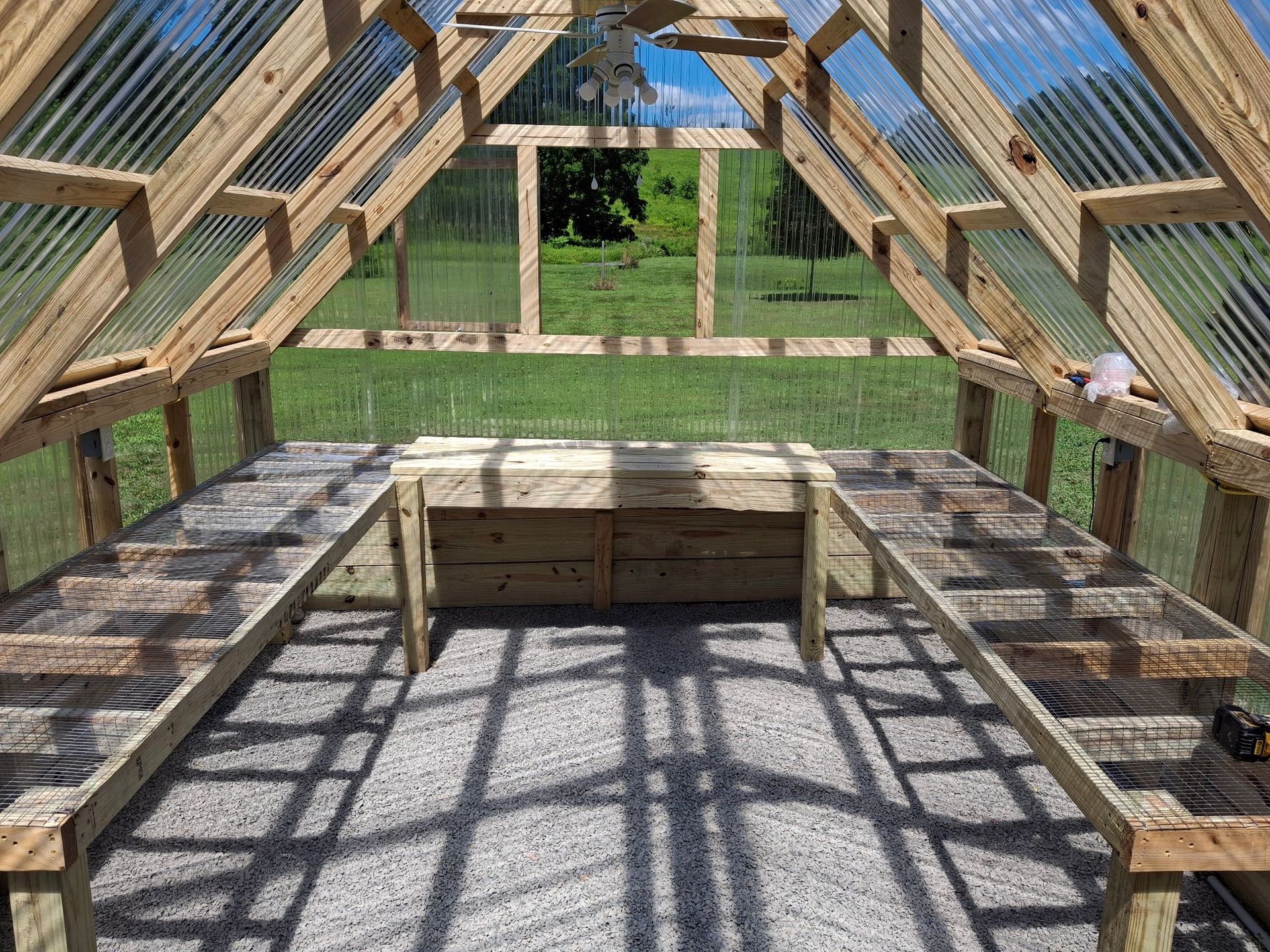 Interior of a wooden greenhouse with transparent roofing. Gravel floor, benches along the sides, and a view of a grassy yard.