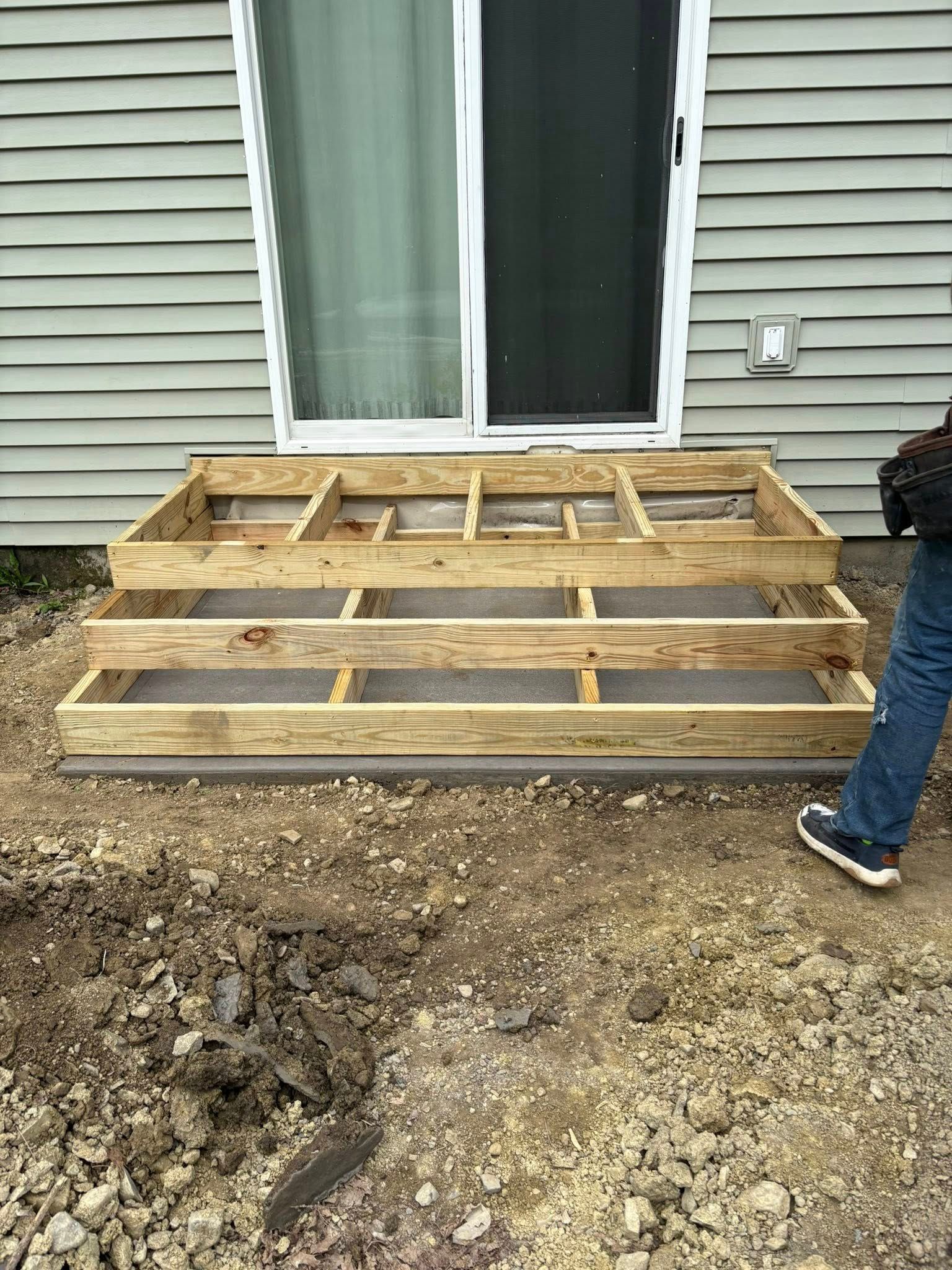 Wooden deck frame construction with three steps in front of a sliding door; a person stands nearby.
