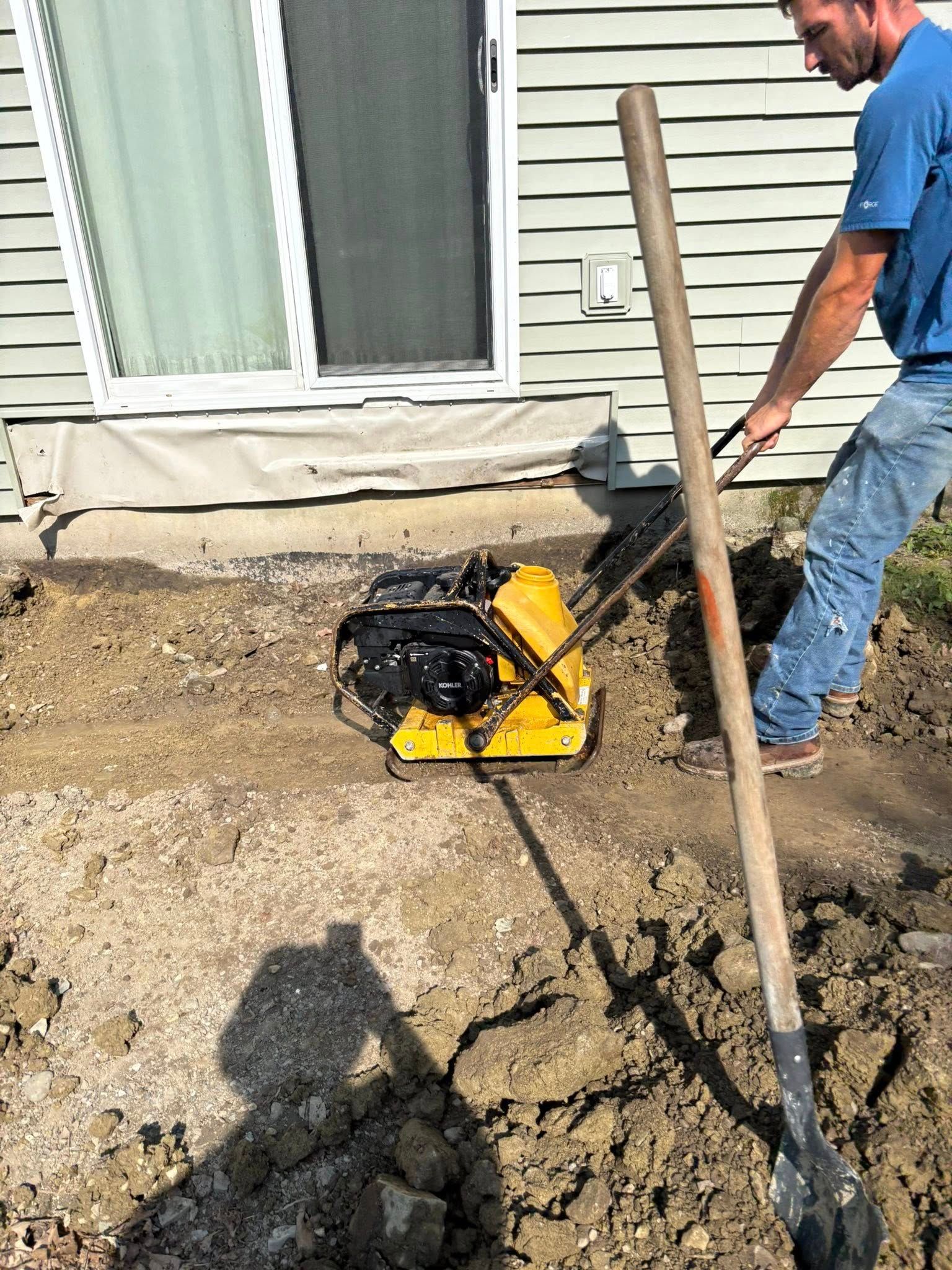 Man using a plate compactor next to a sliding door, compacting gravel base for a patio.