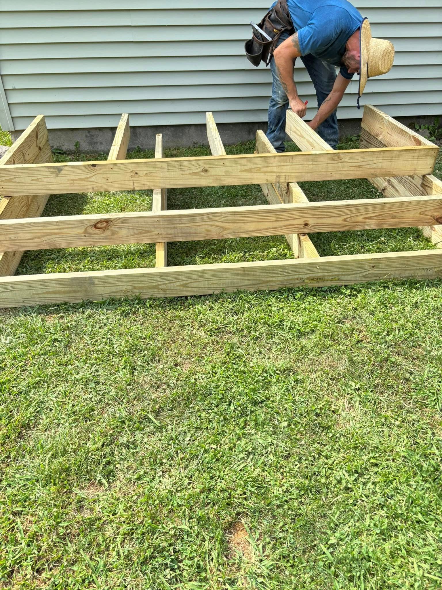 Person building a wooden deck frame on grass next to a house with blue siding.