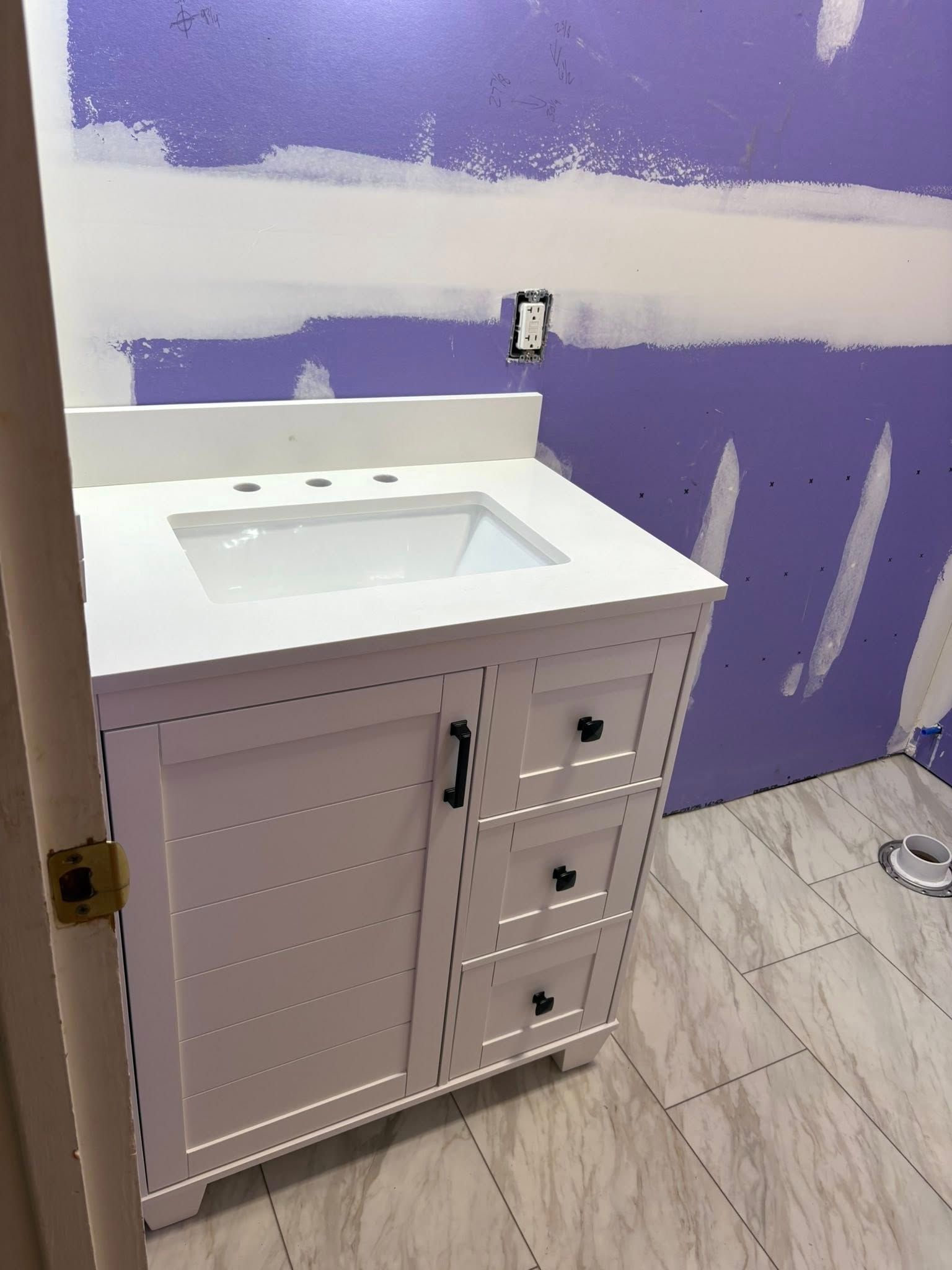 White bathroom vanity with sink and drawers, installed against a purple wall with unfinished drywall.
