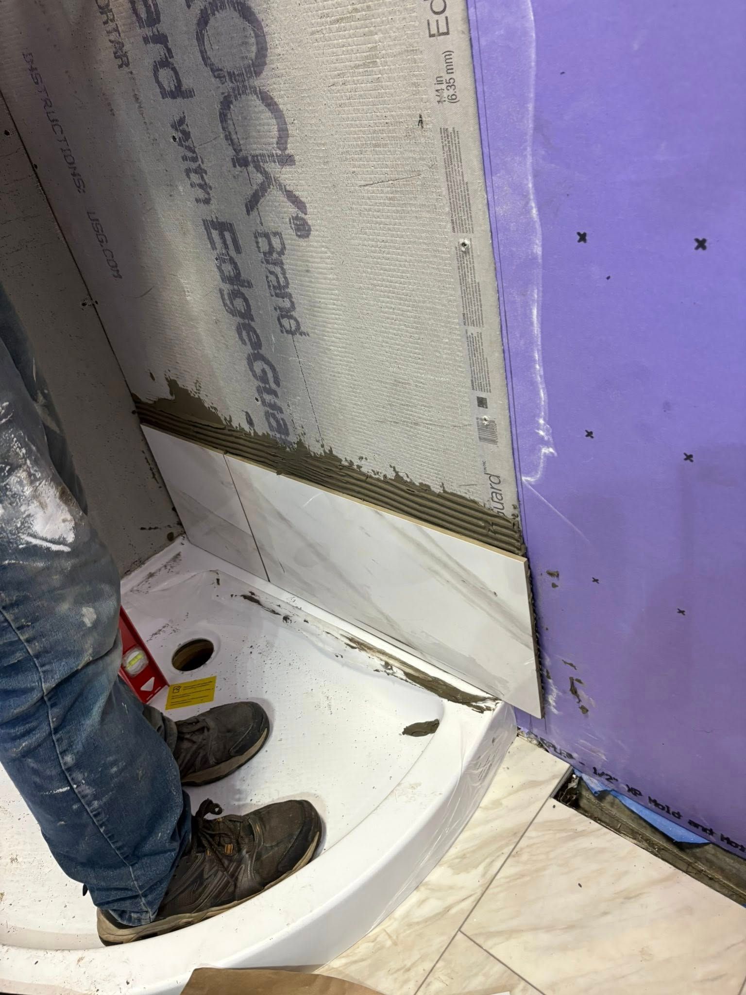 Person installing tile in a shower, using a level. Gray tile, purple wall, and white shower pan.