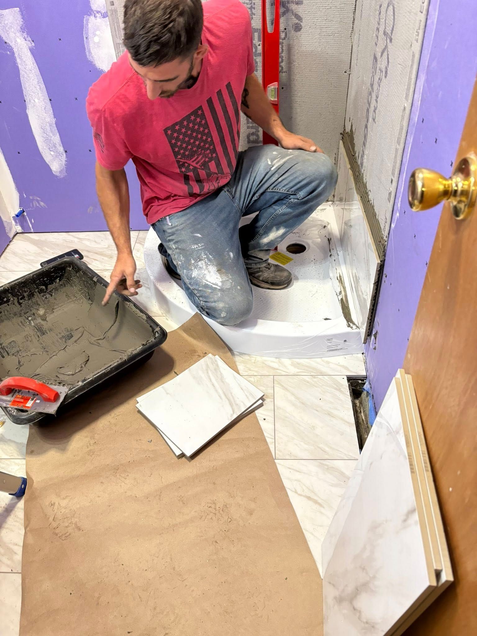 Man kneeling in a bathroom, tiling a shower. Mortar tray and tiles are nearby.