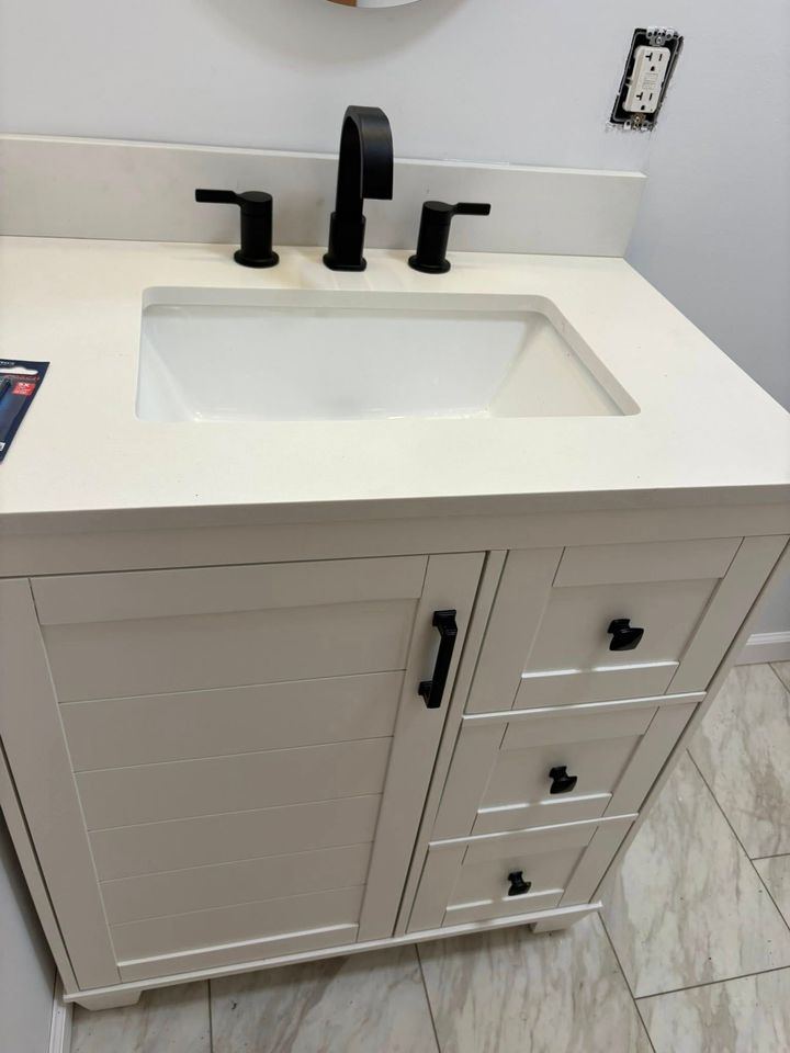 White bathroom vanity with black faucet and hardware; white countertop, sink, and cabinets; tiled floor.